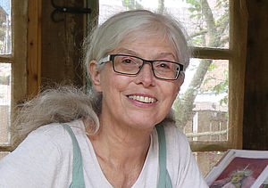 Eine Frau mit langen grauen Haaren und Brille lächelt in die Kamera. Im Hintergrund ein altes Fensterr mir Blick ins Grüne. mi